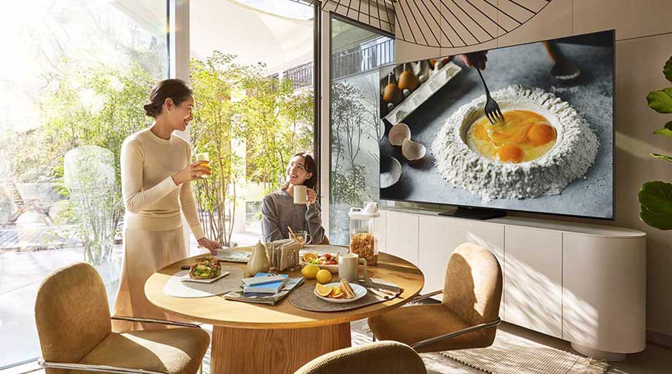 Twee mensen ontbijten aan een ronde tafel met fruit, brood en sap. Op de Samsung-tv staat een kookprogramma met eieren en bloem.