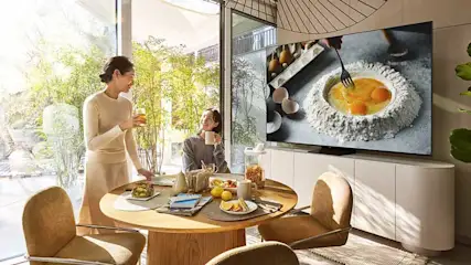 Deux personnes prennent le petit-déjeuner à une table ronde avec fruits, pain et jus. Une TV Samsung diffuse une émission de cuisine avec des œufs.