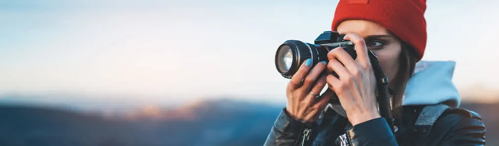 Une personne prend une photo avec un appareil photo noir. Le ciel est bleu clair avec des montagnes au loin.