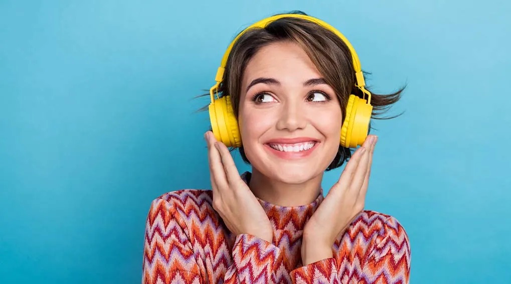 Une femme portant un casque jaune, mains levées sur fond bleu. Elle porte un pull à motifs orange, blanc et rouge.
