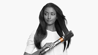 Een close-up van een goudkleurige Dyson Airwrap styler die gebruikt wordt op lang donker haar, tegen een witte achtergrond.