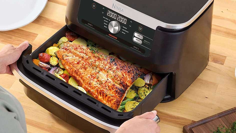 An air fryer on a wooden surface has a drawer open, revealing cooked salmon and vegetables inside.