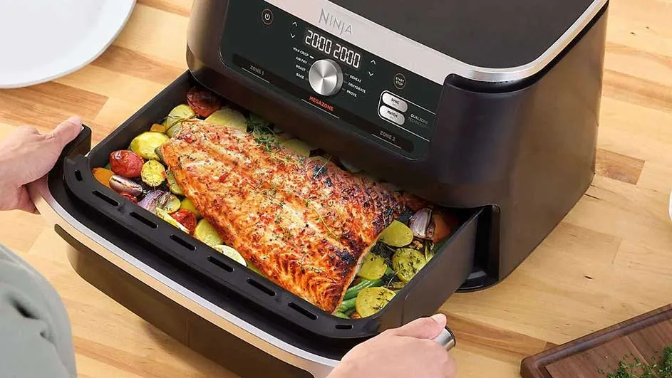 An air fryer on a wooden surface has a drawer open, revealing cooked salmon and vegetables inside.