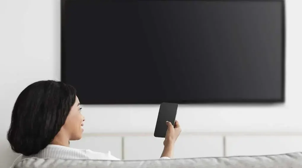 A black TV is mounted on a white wall. A hand holds a black phone. A grey sofa is at the bottom.