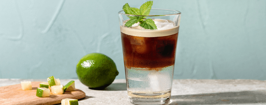 Un café glacé avec des glaçons et de la menthe dans un verre, à côté d’un citron vert et de morceaux de citron vert sur fond bleu-vert clair.