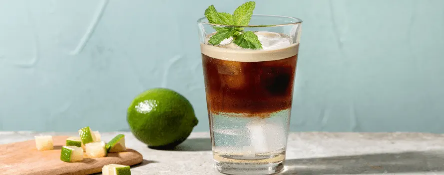 Un café glacé avec des glaçons et de la menthe dans un verre, à côté d’un citron vert et de morceaux de citron vert sur fond bleu-vert clair.