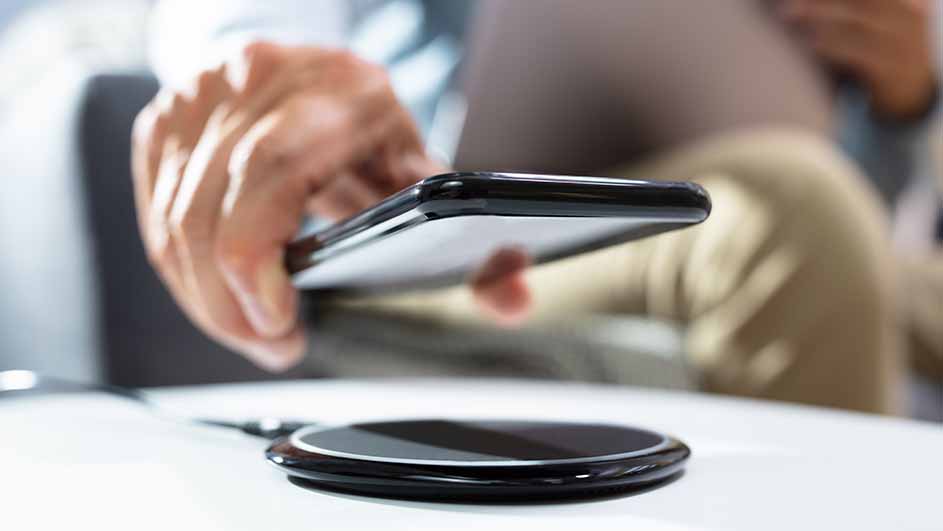 Une main tient un smartphone noir au-dessus d'un chargeur sans fil rond sur une table blanche.