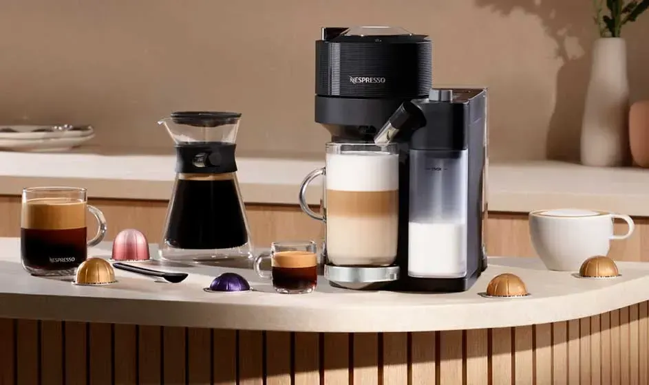 Une machine Nespresso avec réservoir à lait est posée sur une table, entourée de tasses, cafés, capsules et décor beige doux.