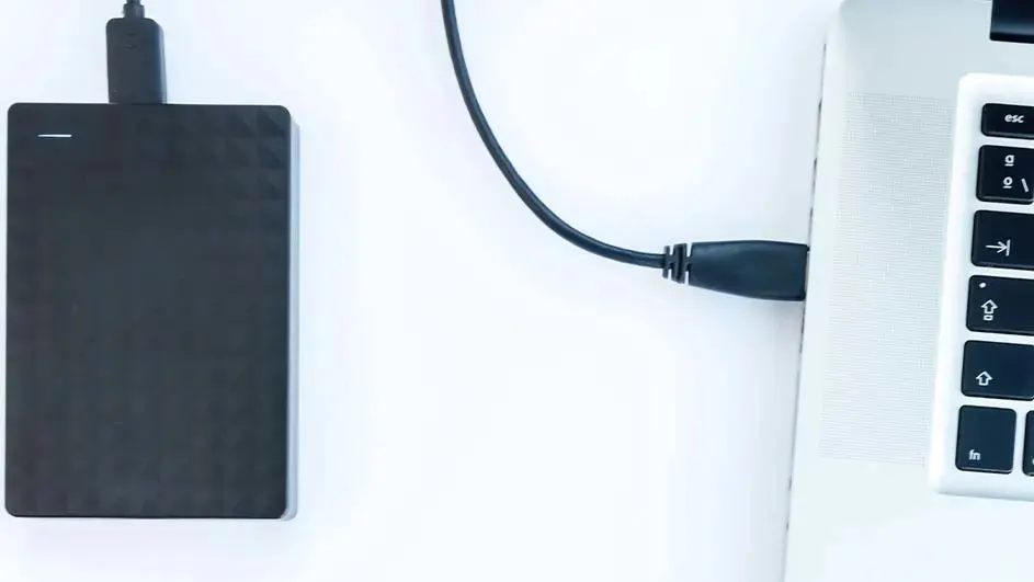 Un disque dur noir est connecté à un ordinateur portable blanc par un câble noir sur une surface blanche.
