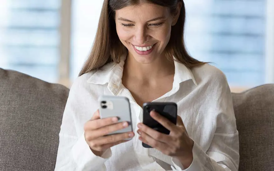 Smiling woman sits on a sofa holding two smartphones, comparing the screens as if she is transferring data from one device to the other.