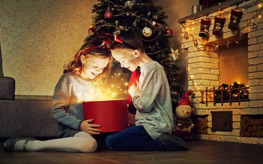 Twee kinderen zitten bij de kerstboom en open een ronde doos vol fonkelend licht, terwijl haardvuur en slingers voor een warme sfeer zorgen.