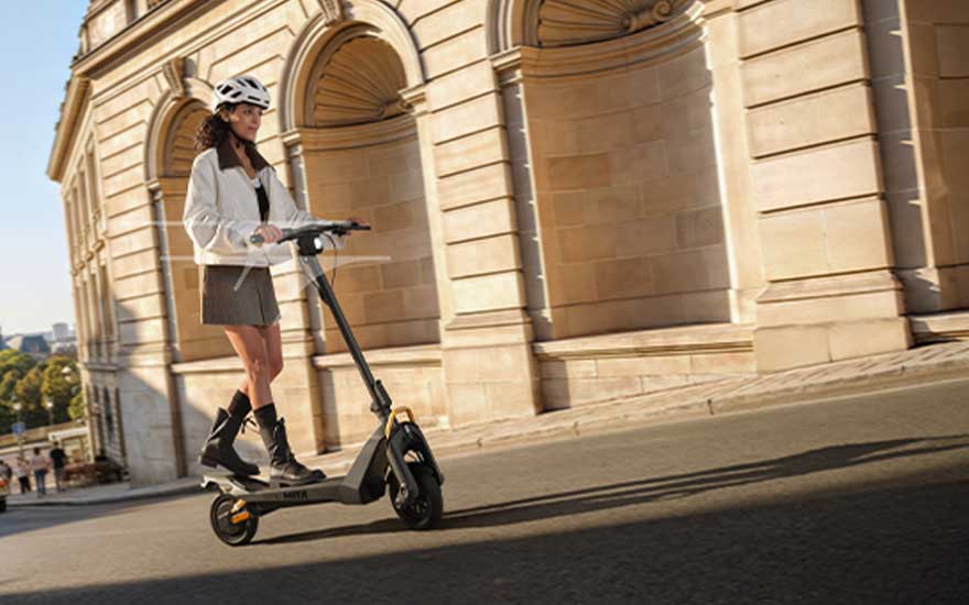 Een vrouw met helm rijdt op een elektrische step langs een historisch gebouw met bogen, in zacht zonlicht op een brede straat.