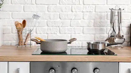Sur un comptoir en bois, une plaque de cuisson avec casserole et marmite, ustensiles de cuisine, dosseret en briques blanches.