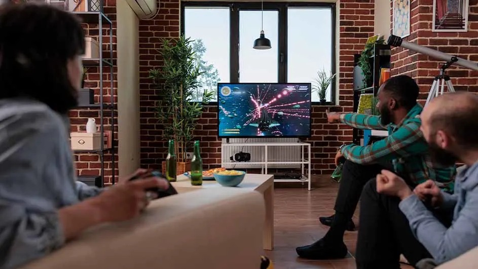 Woonkamer met bakstenen muren en groot raam. Tv toont computerspel. Tafel met bier en snacks.