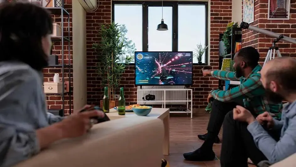 Woonkamer met bakstenen muren en groot raam. Tv toont computerspel. Tafel met bier en snacks.