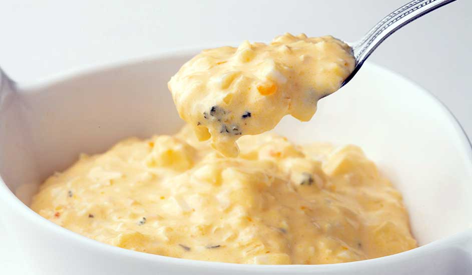 Een zilveren lepel tilt een schep romige, gele aardappelsalade uit een witte kom.