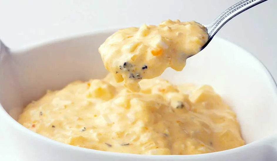 Een zilveren lepel tilt een schep romige, gele aardappelsalade uit een witte kom.