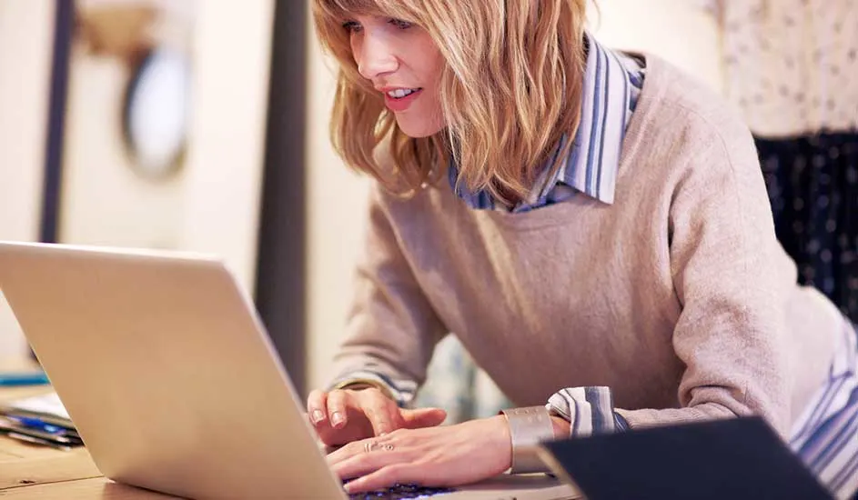 Een laptop staat op een houten tafel, met op de achtergrond een vage kamer en een gedeelte van een blauw gestreept hemd.