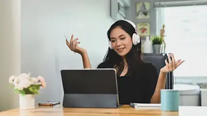 Een laptop op een houten bureau, met een blauwe beker, bloemen en een telefoon, tegen een witte achtergrond.