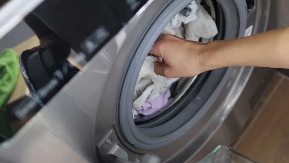 Een hand reikt in een zilverkleurige wasmachine gevuld met witte en paarse kleding.