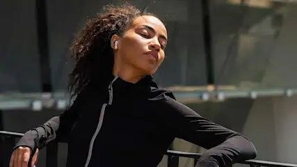 A person in a black jacket with a white earbud rests on a black railing. A building is in the background.