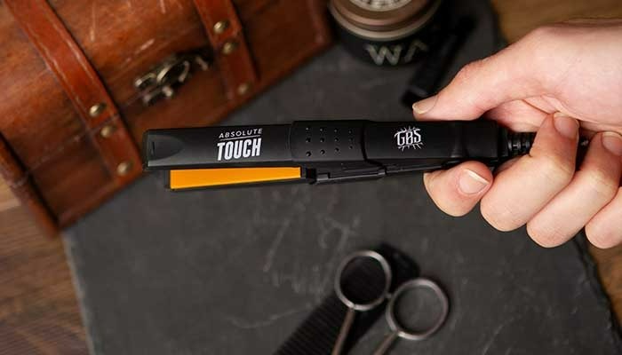 A hand holds a black hair straightener with orange plates, labeled "Absolute Touch GBS," on a dark surface with scissors and a brown chest.