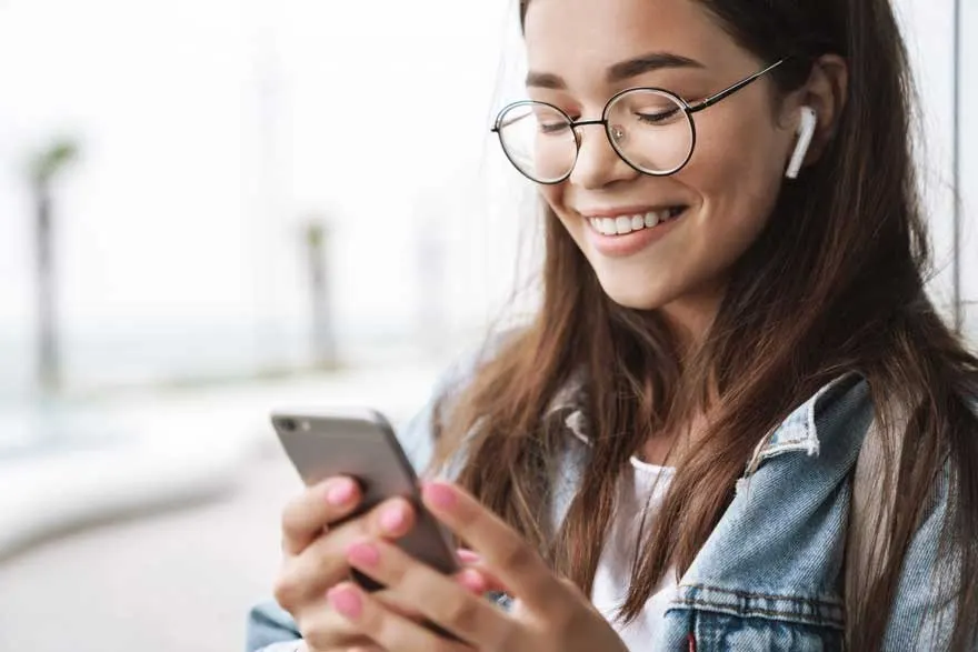 Une personne tient un smartphone gris. Elle porte des lunettes rondes et une veste en jean bleue. Des écouteurs blancs sont visibles.