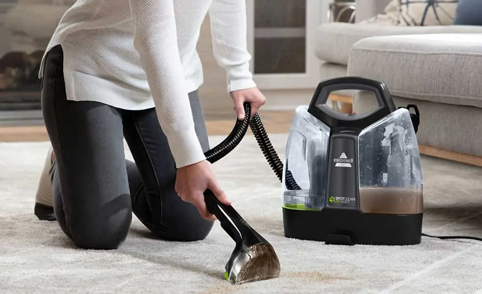Aspirateur noir sur tapis beige à motifs, avec réservoirs transparents. Un tuyau noir est connecté. Canapé gris clair en arrière-plan.