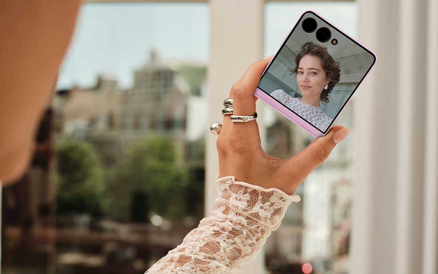 Een kleine opvouwbare Galaxy Z Flip8 toont een selfieportret op het buitenscherm, vastgehouden bij een raam met stadszicht.