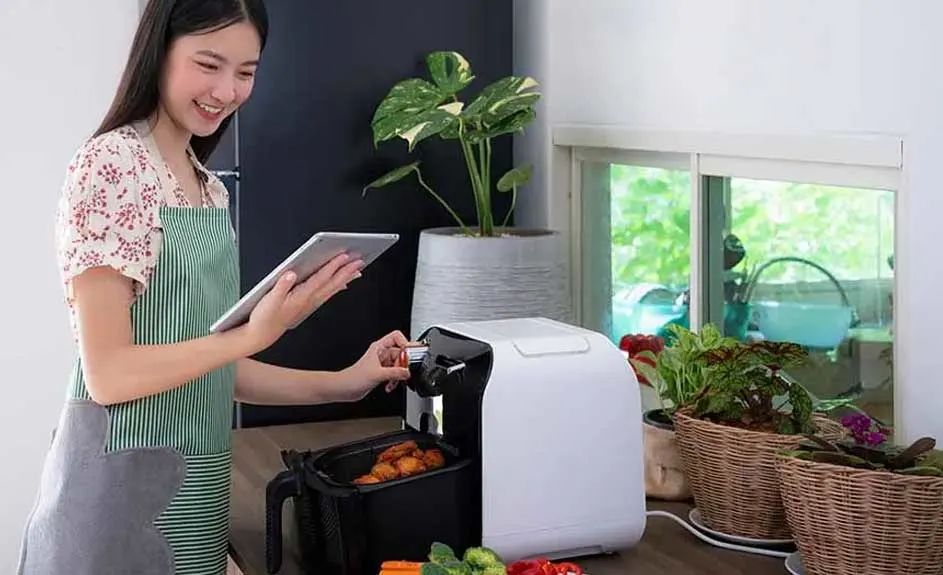Een witte airfryer op een aanrecht met groenten. Een tablet en planten zijn ook zichtbaar.