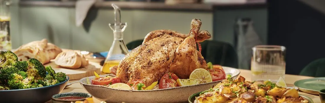 Roast chicken on a platter with tomatoes and lemon slices, plus sides of broccoli, bread, and potatoes on a table.