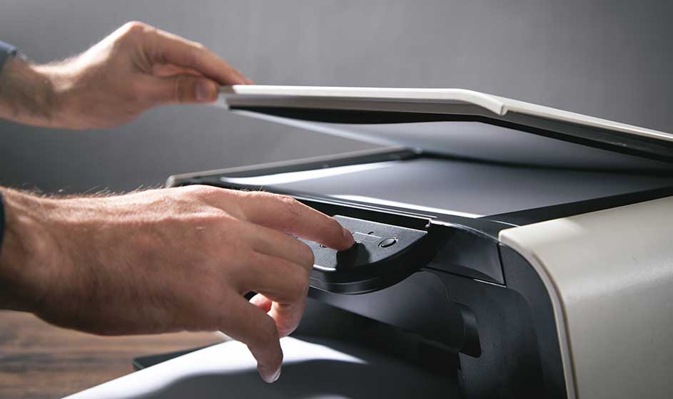 Handen bedienen een printer of scanner met geopend deksel, terwijl een vel papier op de glasplaat ligt in een moderne kantooromgeving.