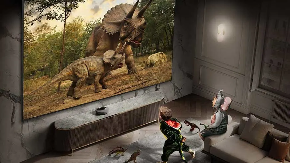 Een woonkamer met een groot scherm waarop een afbeelding van dinosaurussen te zien is, met een console eronder en een beige sofa.