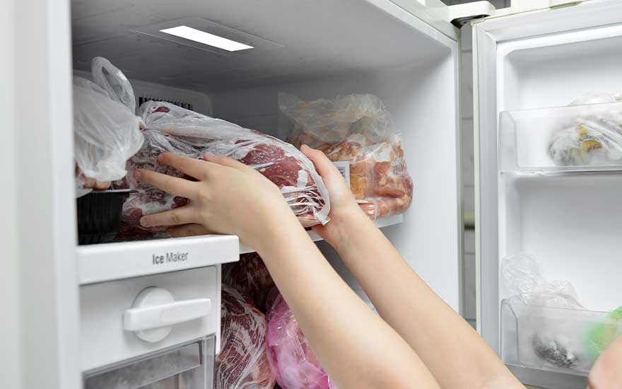 Dans un réfrigérateur blanc, des mains tiennent un sac de viande congelée. D'autres sacs sont visibles à l'intérieur.