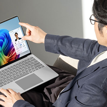 Een grijze laptop toont een videocallinterface. Een vinger raakt het scherm aan. Op de achtergrond is een effen grijze muur te zien.