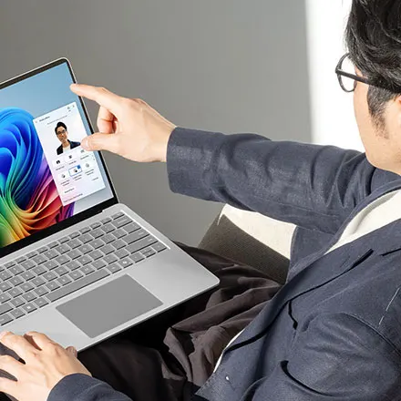 Un ordinateur portable argenté affiche un écran avec un fond d'écran coloré et une fenêtre d'appel vidéo. Un doigt pointe l'écran.