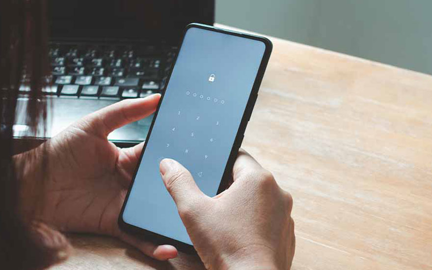 Een hand ontgrendelt een zwarte smartphone met een numeriek slot op een houten tafel, met een laptop op de achtergrond.