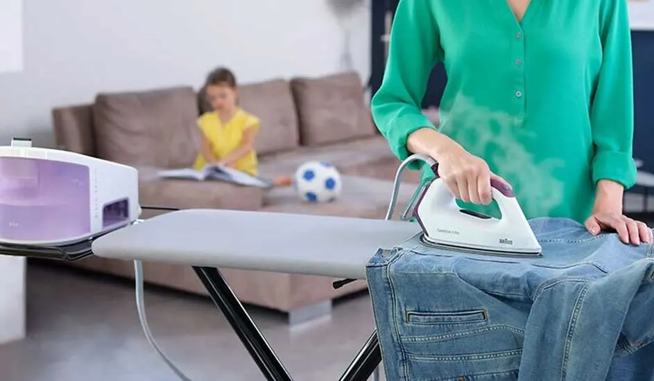 Iemand strijkt een blauw jeansjasje op een strijkplank. Op de achtergrond staat een bruine zetel en een voetbal.