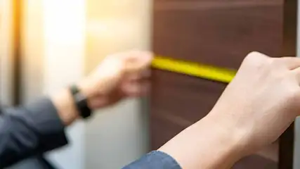 Une personne mesure un panneau brun avec un ruban à mesurer jaune. La main droite tient le ruban, la gauche le fixe.