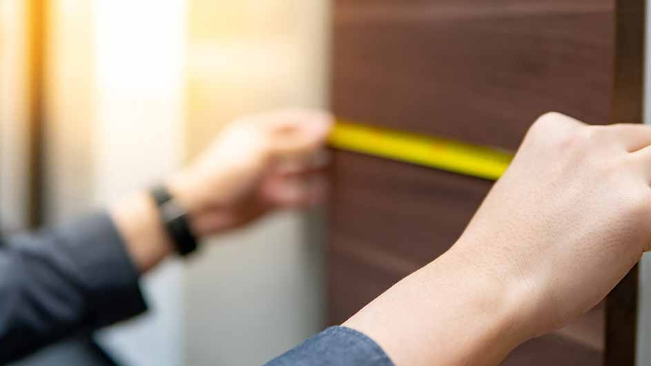 Twee handen meten een bruine houten deur met een geel meetlint. Op de achtergrond is een vage witte muur te zien.