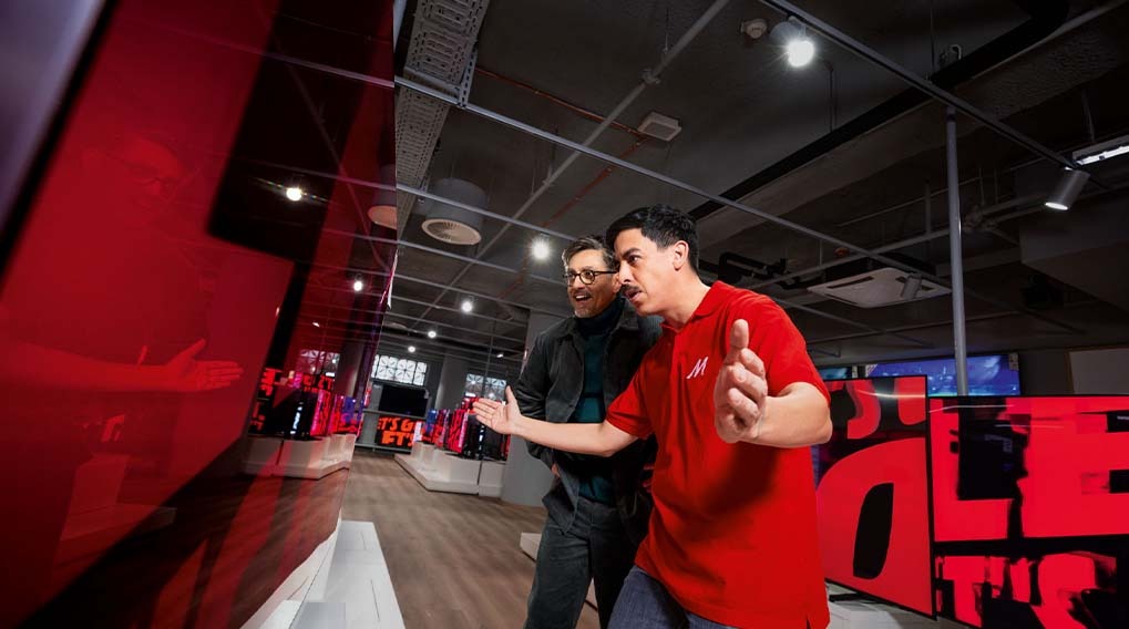 In a retail setting, two individuals view a display of red and black screens under a dropped ceiling with visible ducts and lighting fixtures.