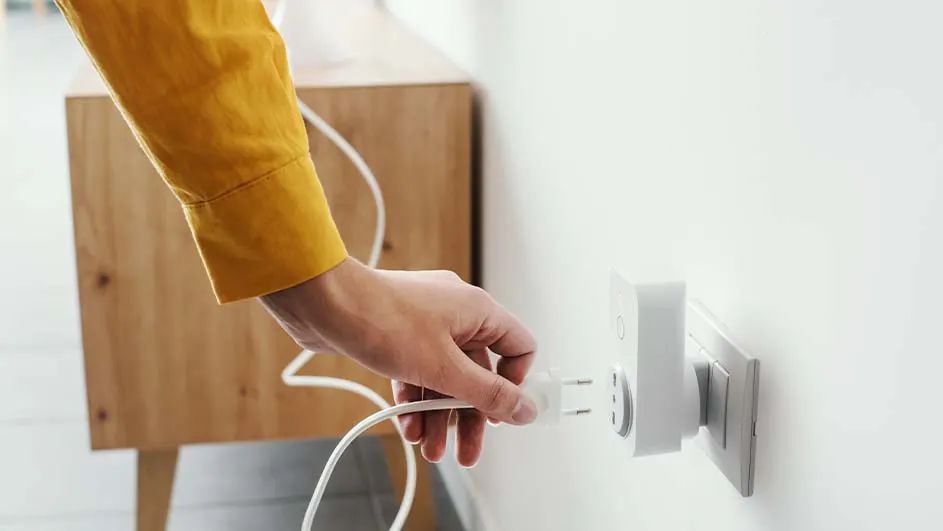 Een witte stekker wordt in een wit stopcontact gestoken tegen een witte muur, met een houten kast op de achtergrond.