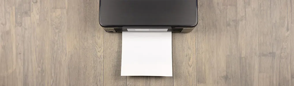 Une imprimante noire imprime une feuille blanche sur un parquet en bois clair.