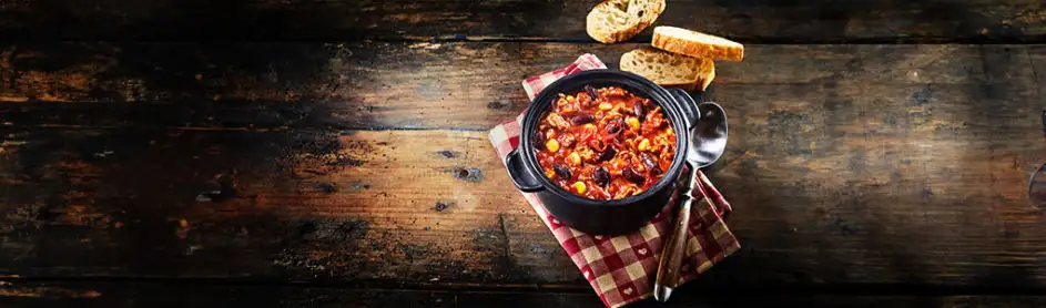 Een zwarte pan met chili staat op een rood-wit geruit doek, naast een lepel en sneetjes brood op een houten tafel.