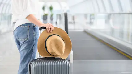 Een bruine hoed ligt op een grijze koffer in een lichte hal met een rollende loopband.