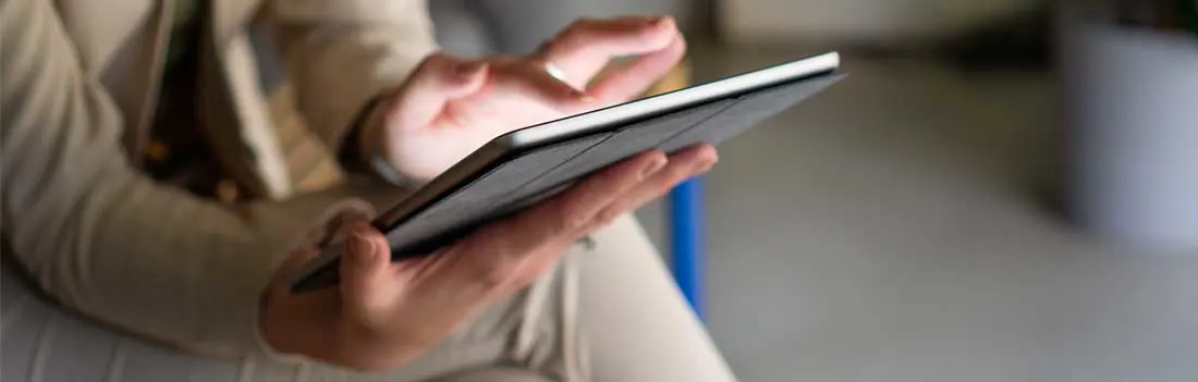 Une personne tient une tablette et touche l’écran, assise dans un intérieur calme aux couleurs neutres et à l’arrière-plan flou.