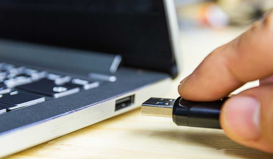 Zwarte USB-stick steekt in een zilverkleurige laptop op een houten tafel. De laptop is open en het toetsenbord is zichtbaar.