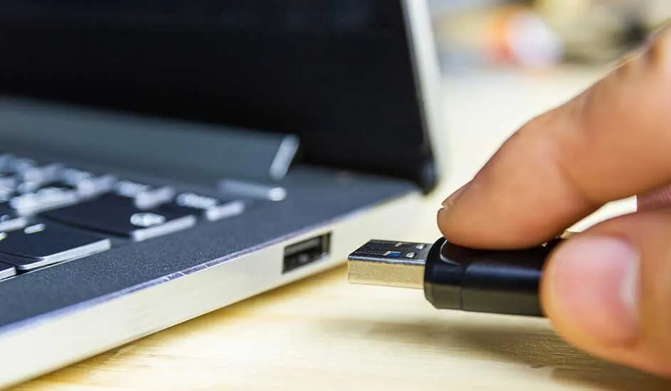 Zwarte USB-stick steekt in een zilverkleurige laptop op een houten tafel. De laptop is open en het toetsenbord is zichtbaar.