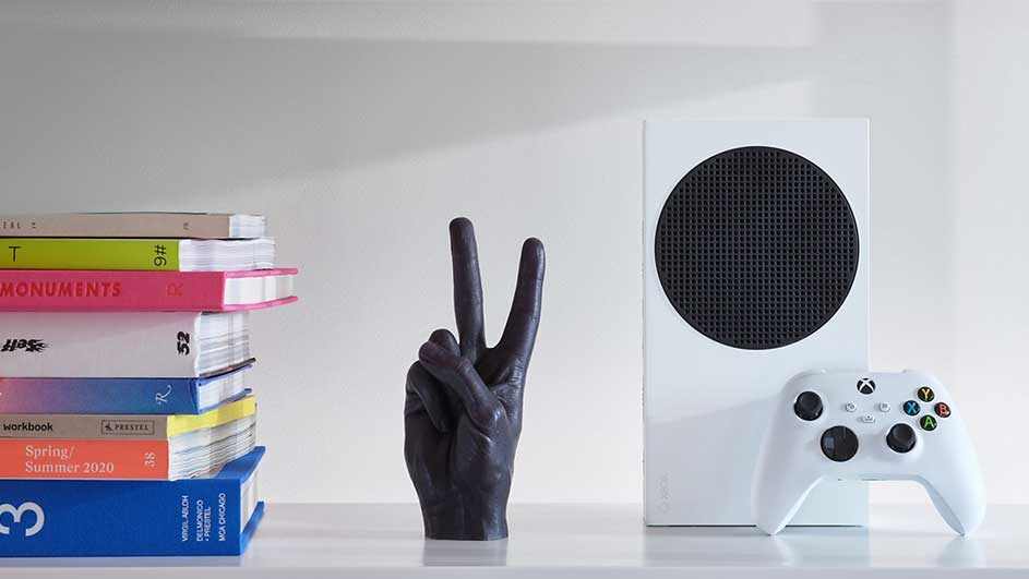 Une console Xbox blanche avec un contrôleur, une main noire en forme de V et une pile de livres colorés sur une étagère blanche.