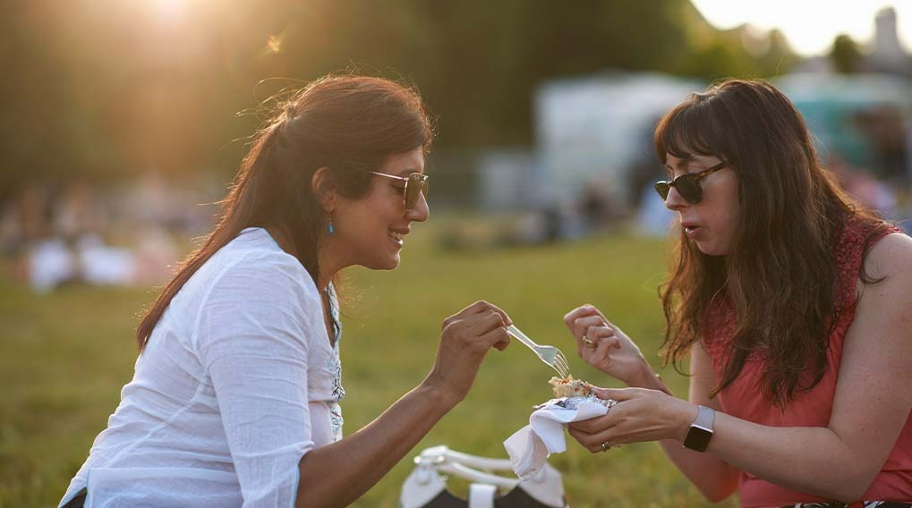 Twee mensen delen eten op een grasveld met bomen en een gebouw op de achtergrond.
